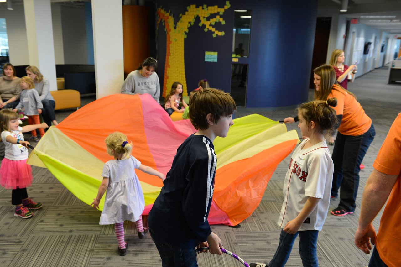 Children and adults play together with a large colorful parachute in an indoor office setting.