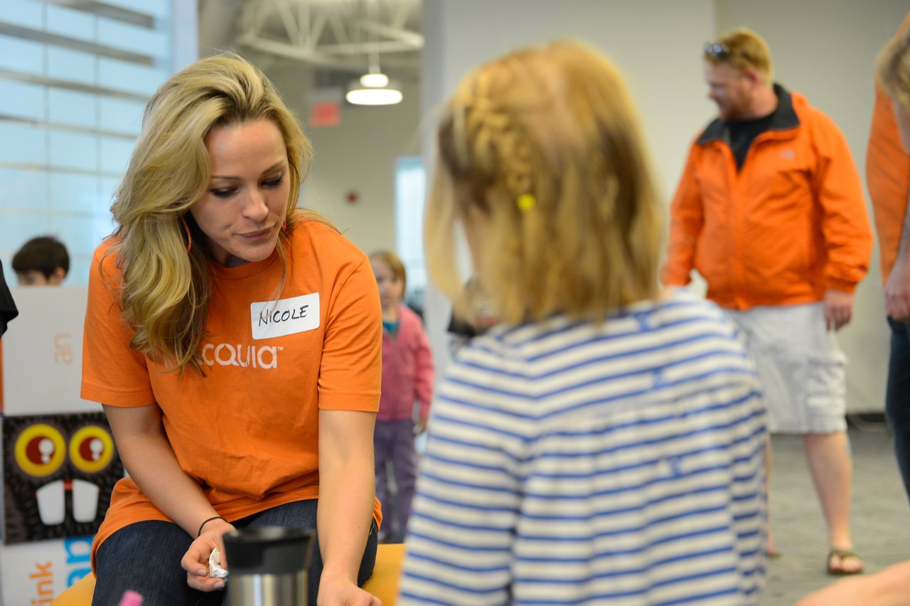 A woman in an orange shirt interacts with a child in a striped shirt at a workplace event.