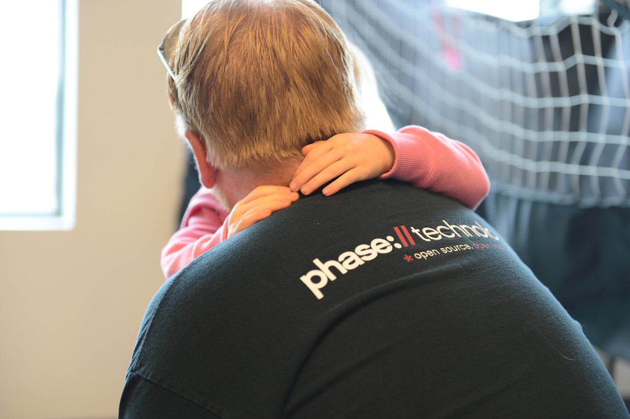 A child in a pink sweater hugs an adult wearing a black "Phase II Technology" shirt from behind.
