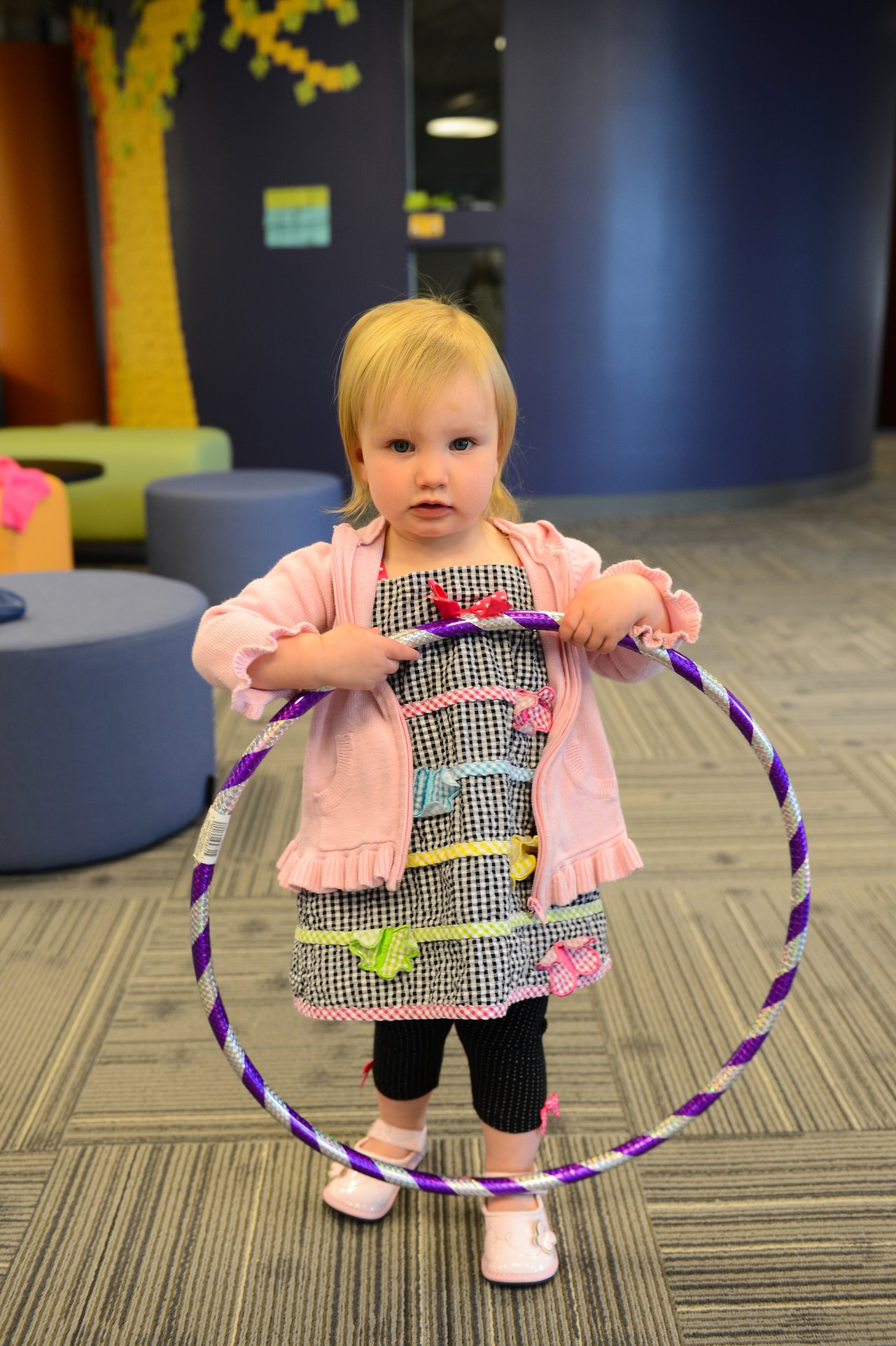 A young child holds a purple hula hoop while standing in an office space during a family event.