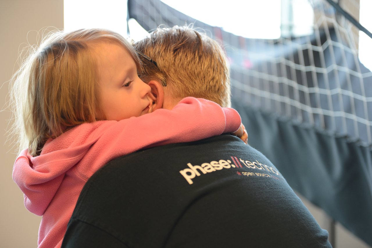 A young child in a pink hoodie hugs an adult wearing a black shirt at a workplace event.