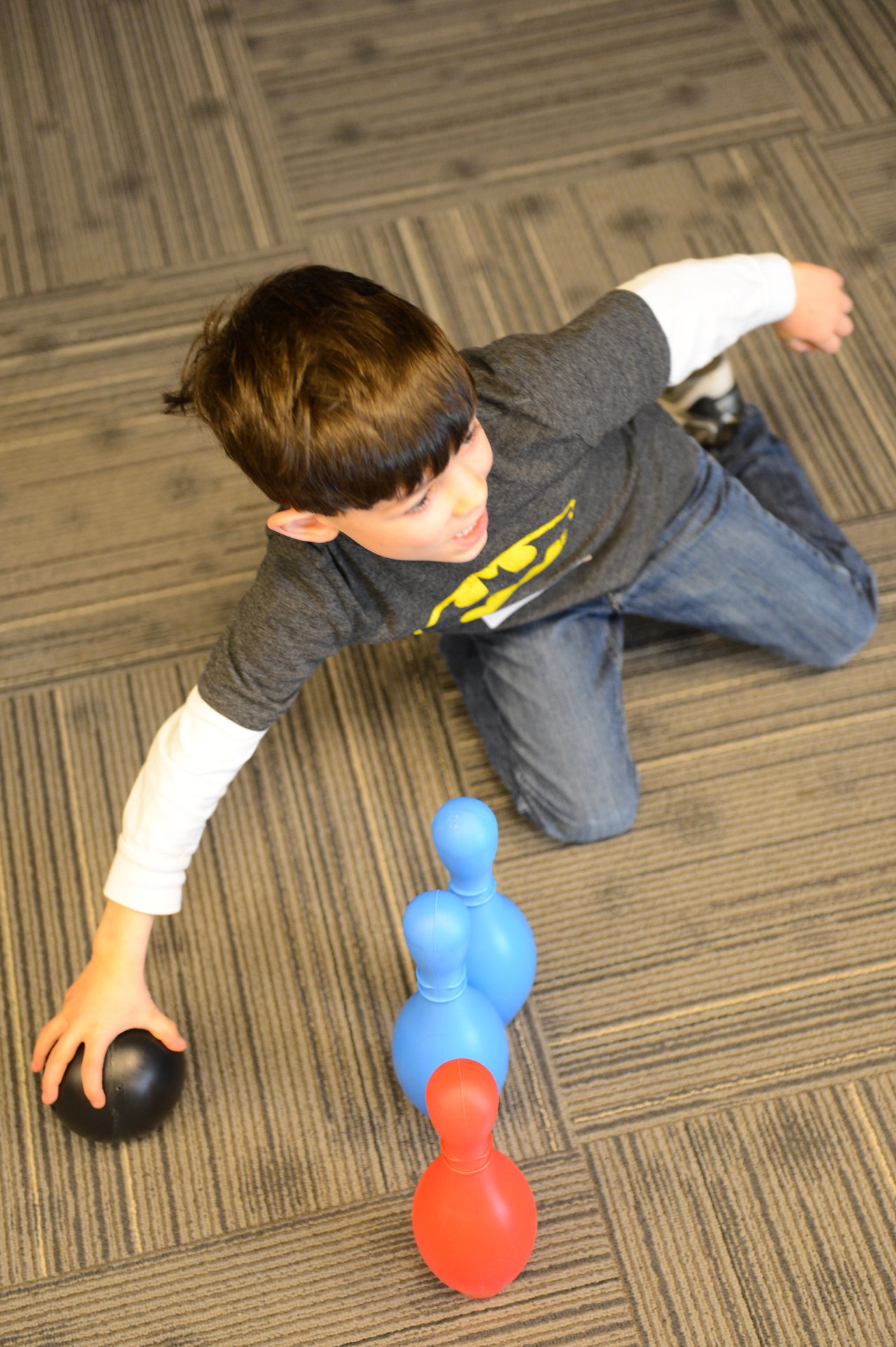 A child in a Batman shirt prepares to roll a small black ball toward colorful plastic bowling pins.