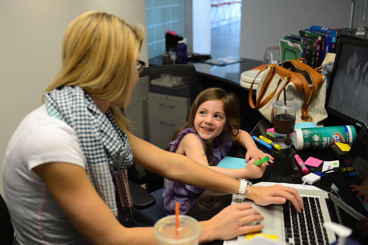 A woman types on a laptop while a young girl smiles and colors with markers at a desk.