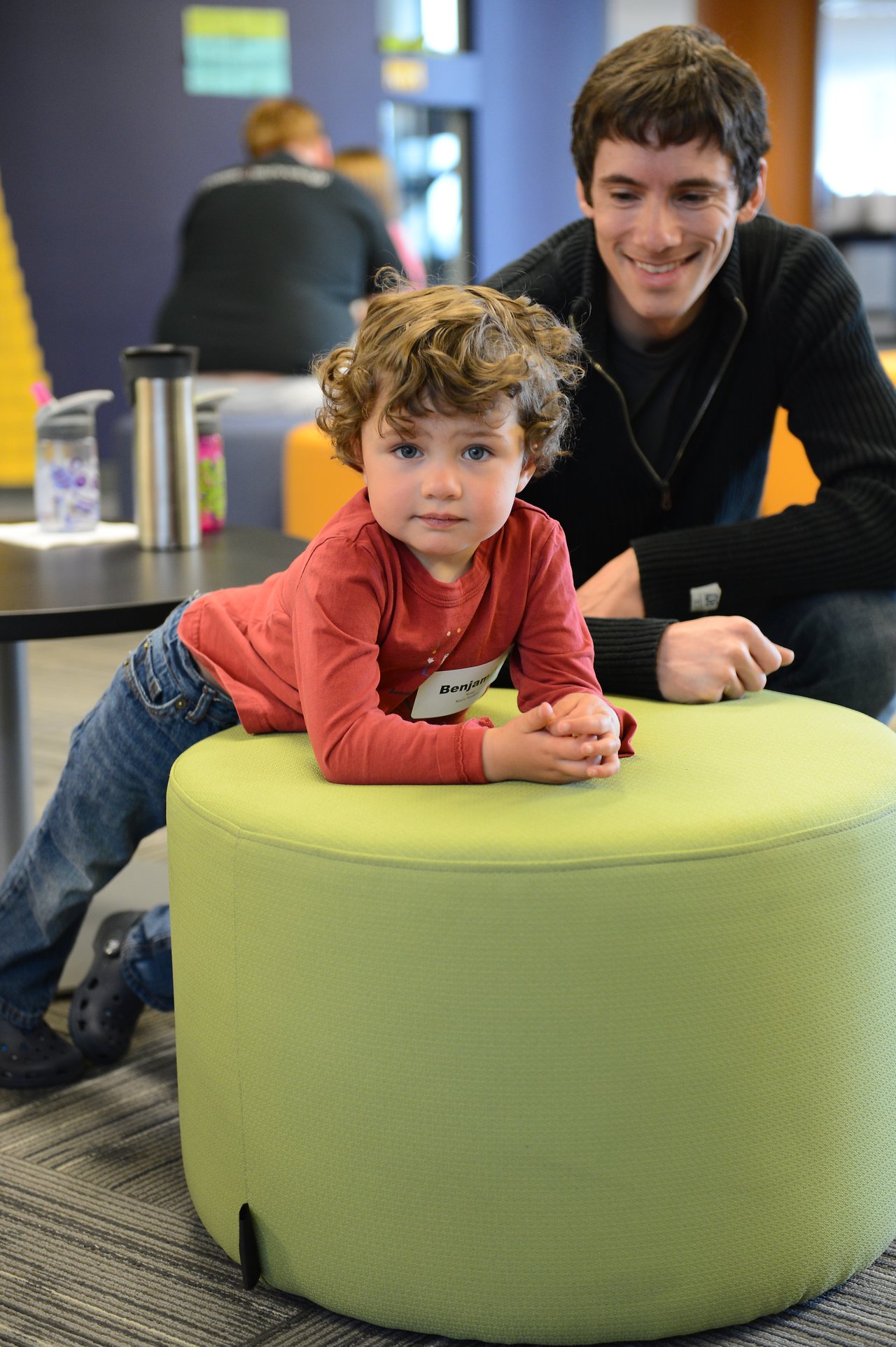 A young child leans on a green ottoman while an adult nearby smiles and watches.