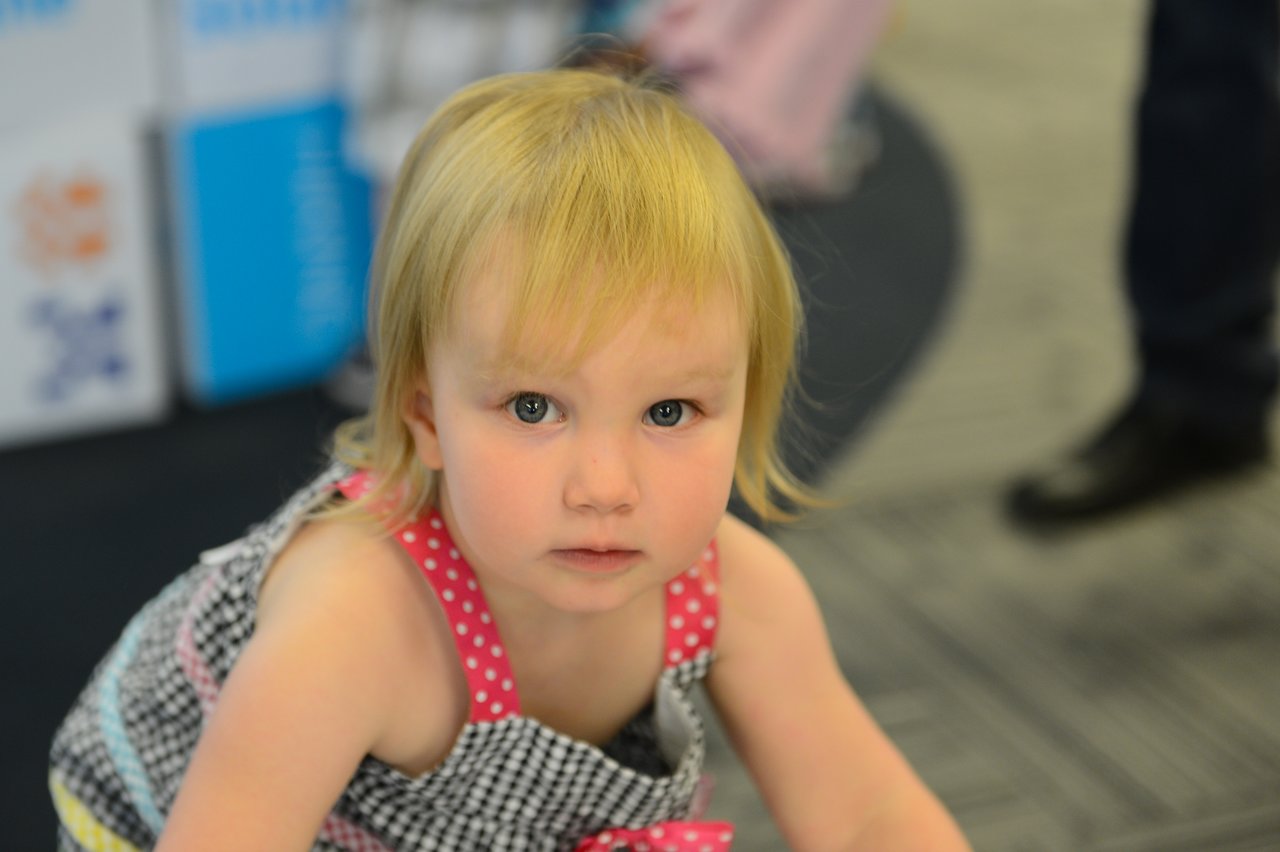 A young child with blonde hair looks directly at the camera while crouching indoors.