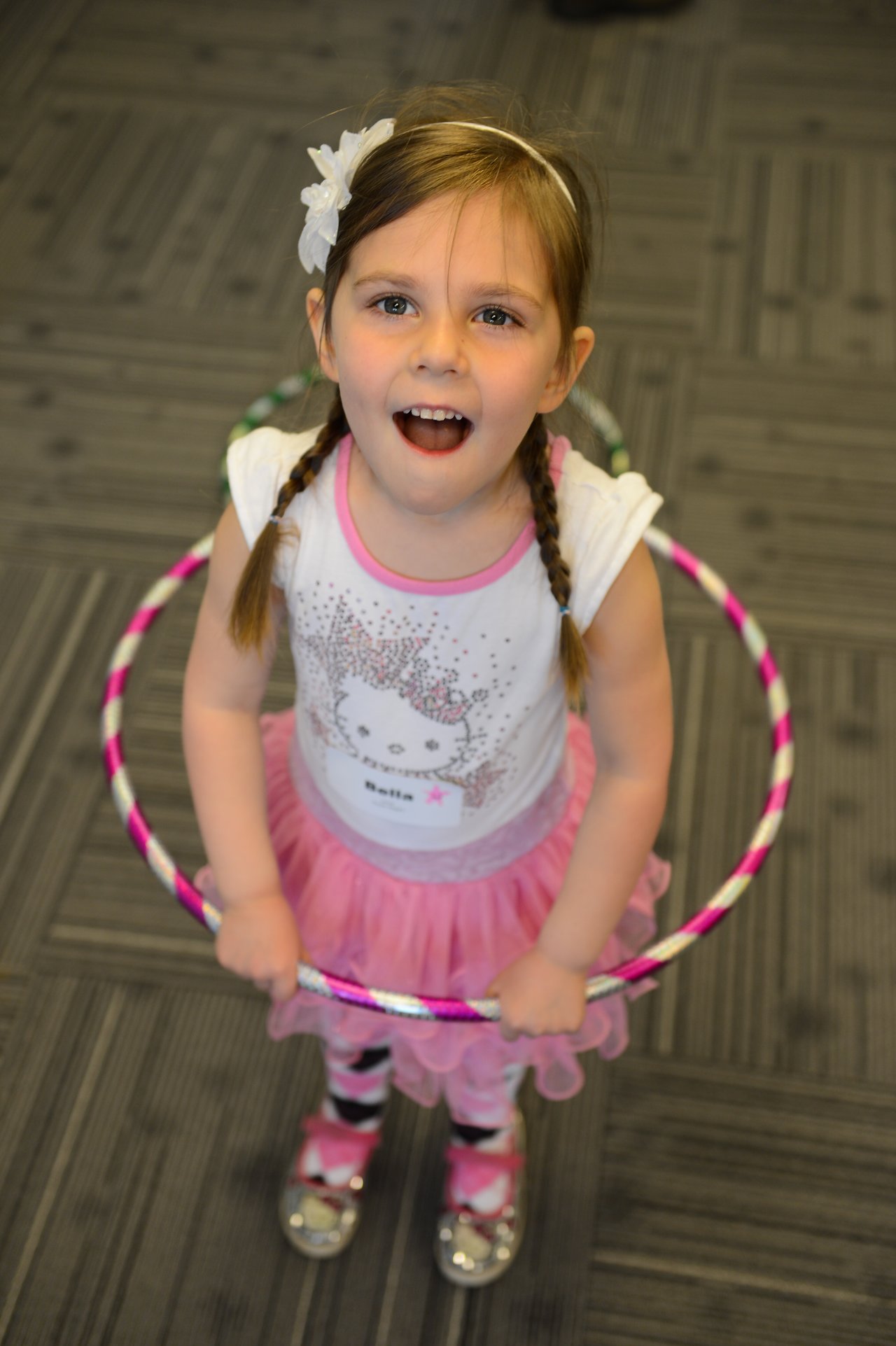 A young girl in a pink outfit holds a hula hoop and looks up with an excited expression.