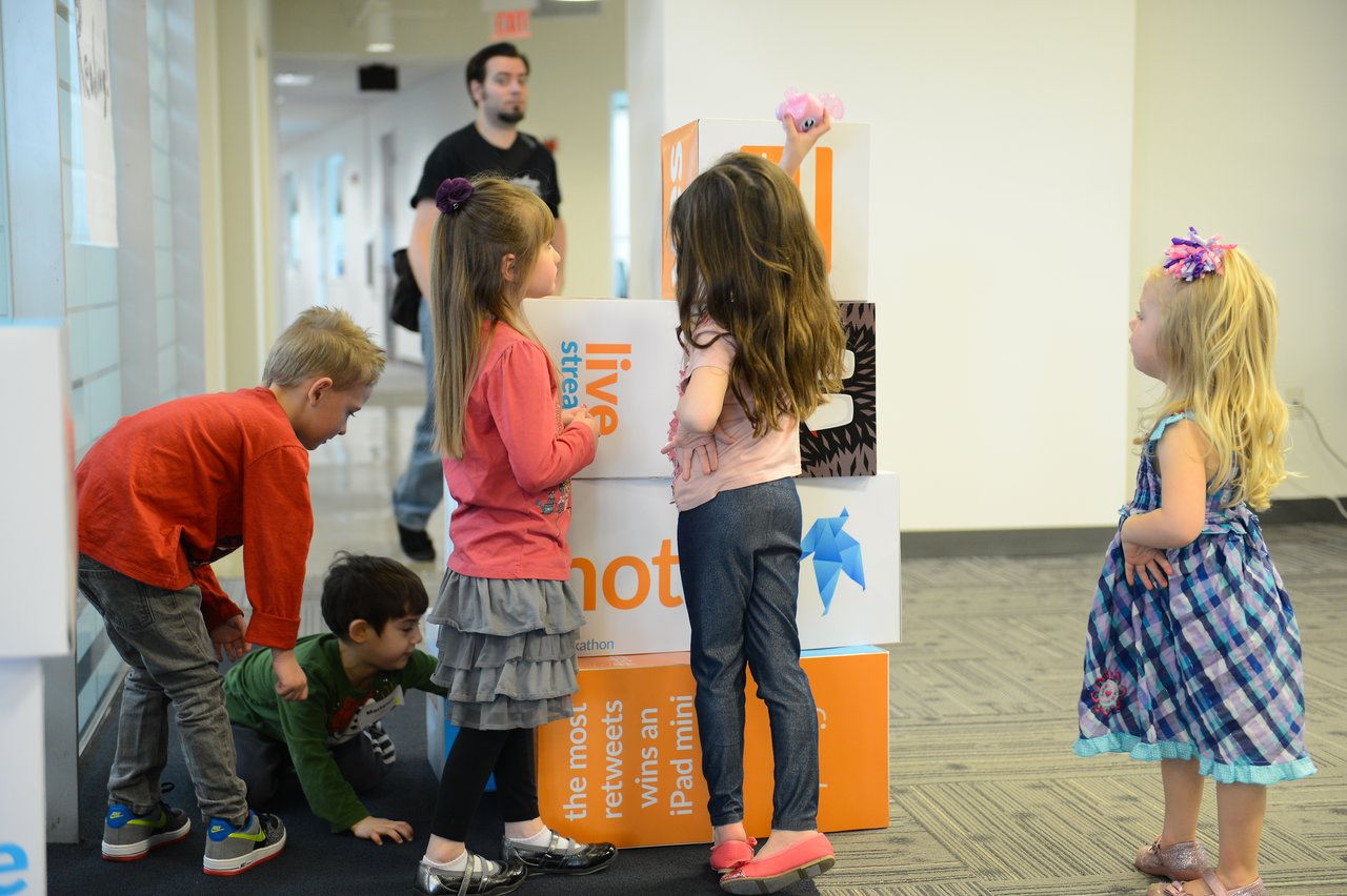 Several children gather around stacked boxes, with one reaching to place a small toy on top.