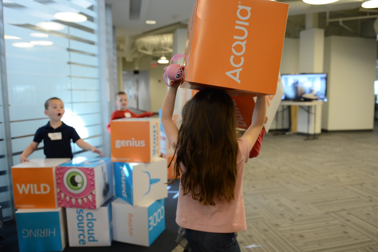 Children play with large colorful boxes in an office, with one child lifting an "Acquia" box over their head.