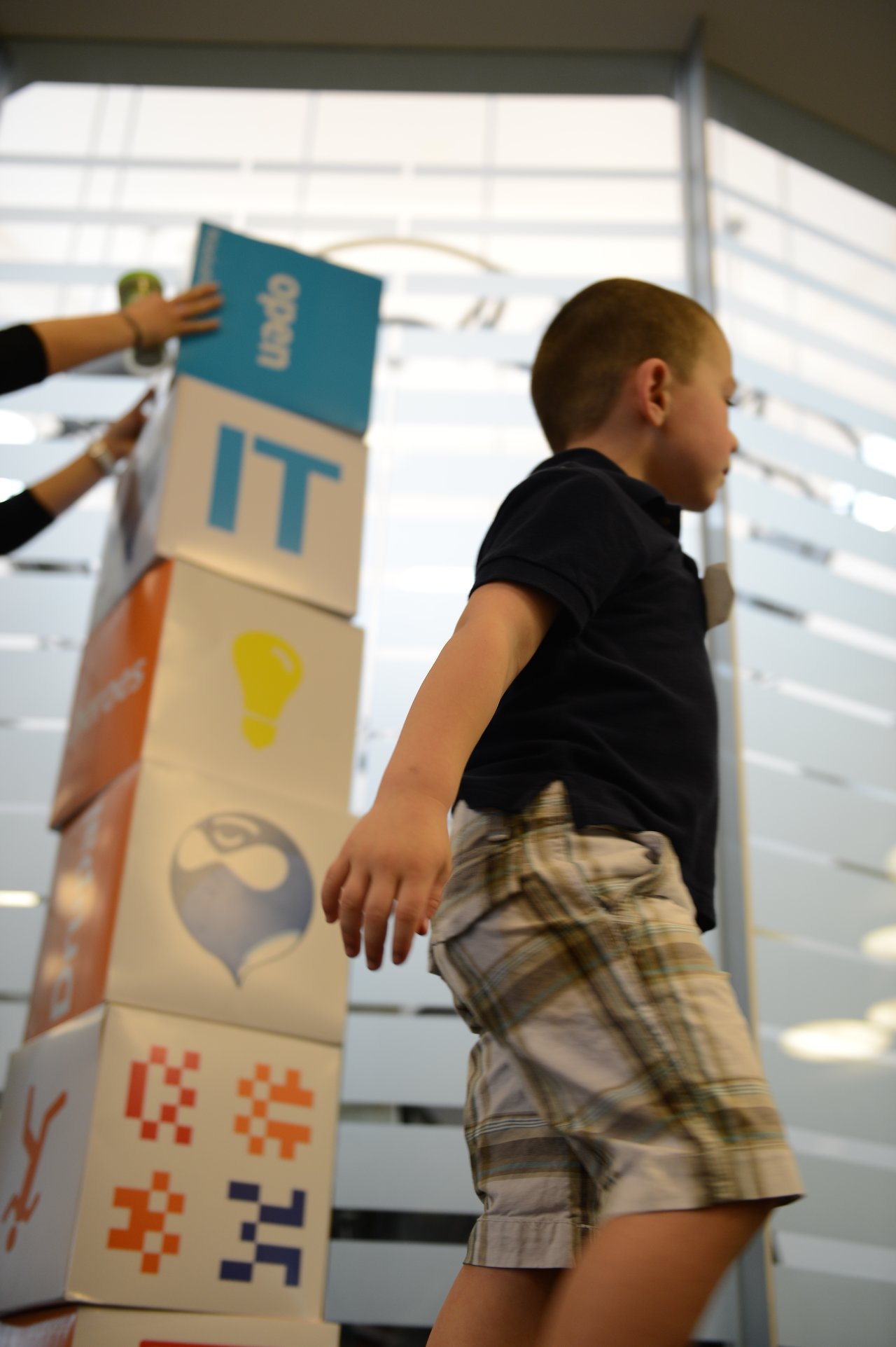 A child walks past a tall stack of branded blocks while an adult adjusts the top one.