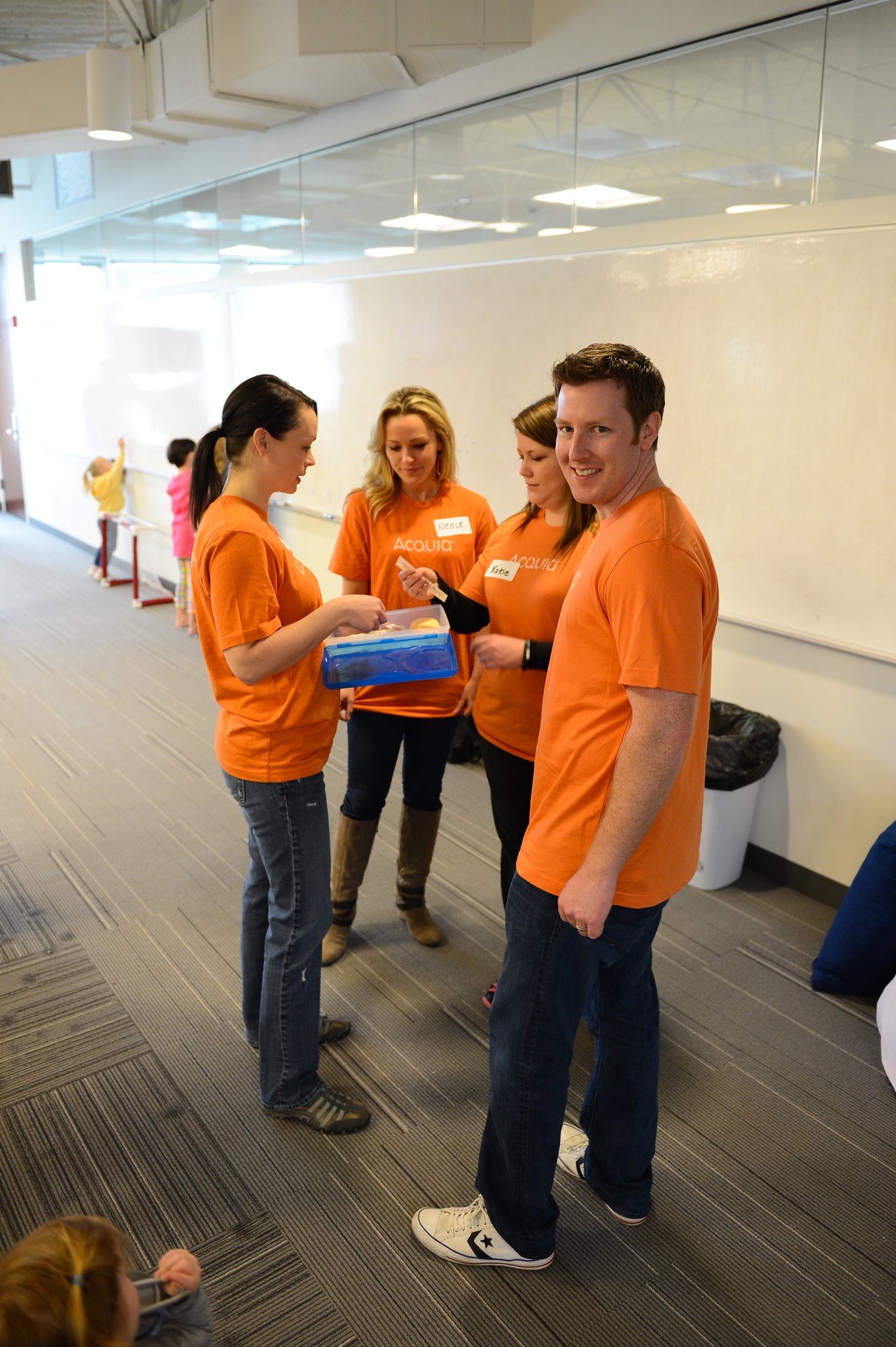 Four people in matching orange shirts stand together, holding and discussing a blue container in an office setting.