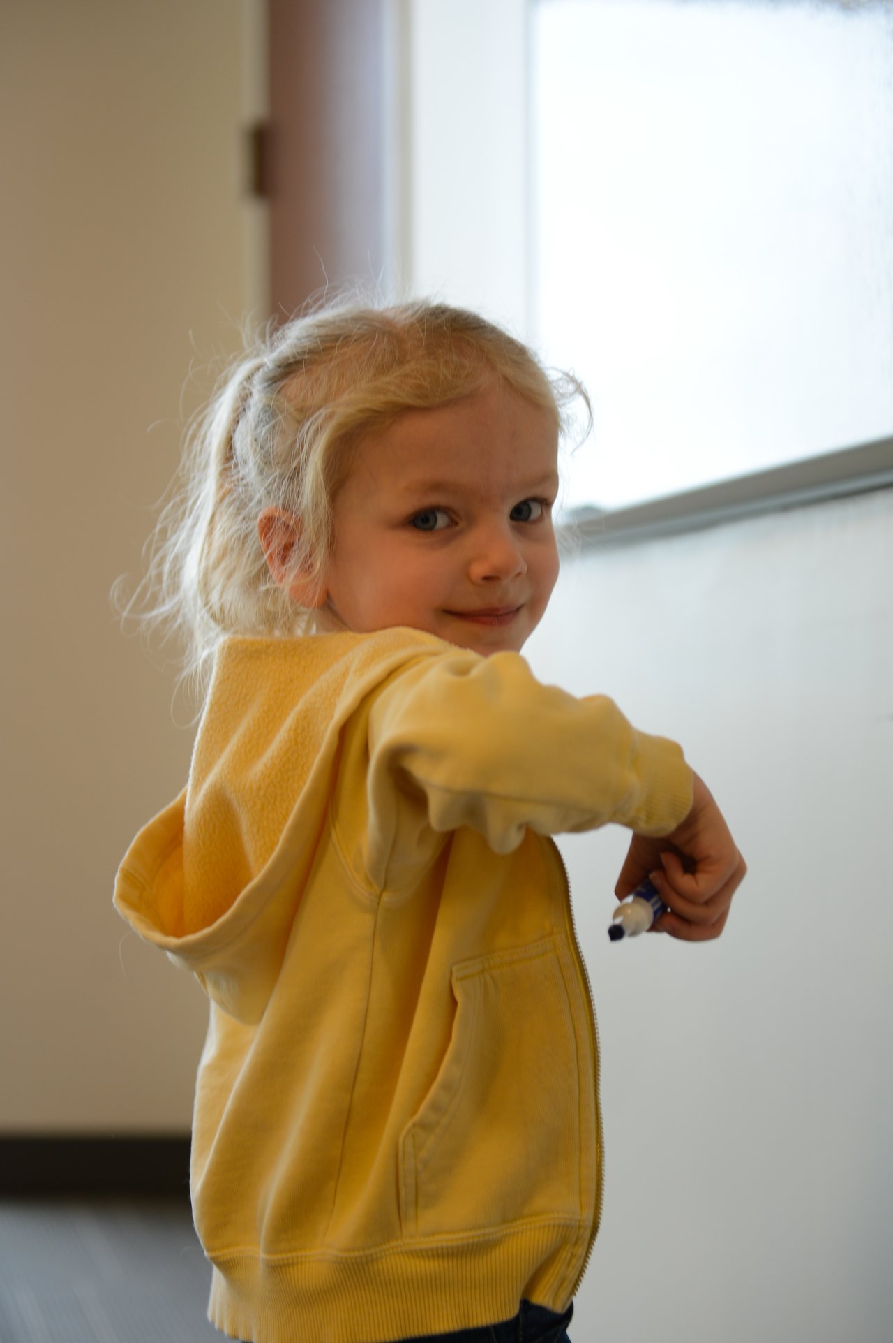 A young child in a yellow hoodie holds a marker and looks back while standing near a whiteboard.