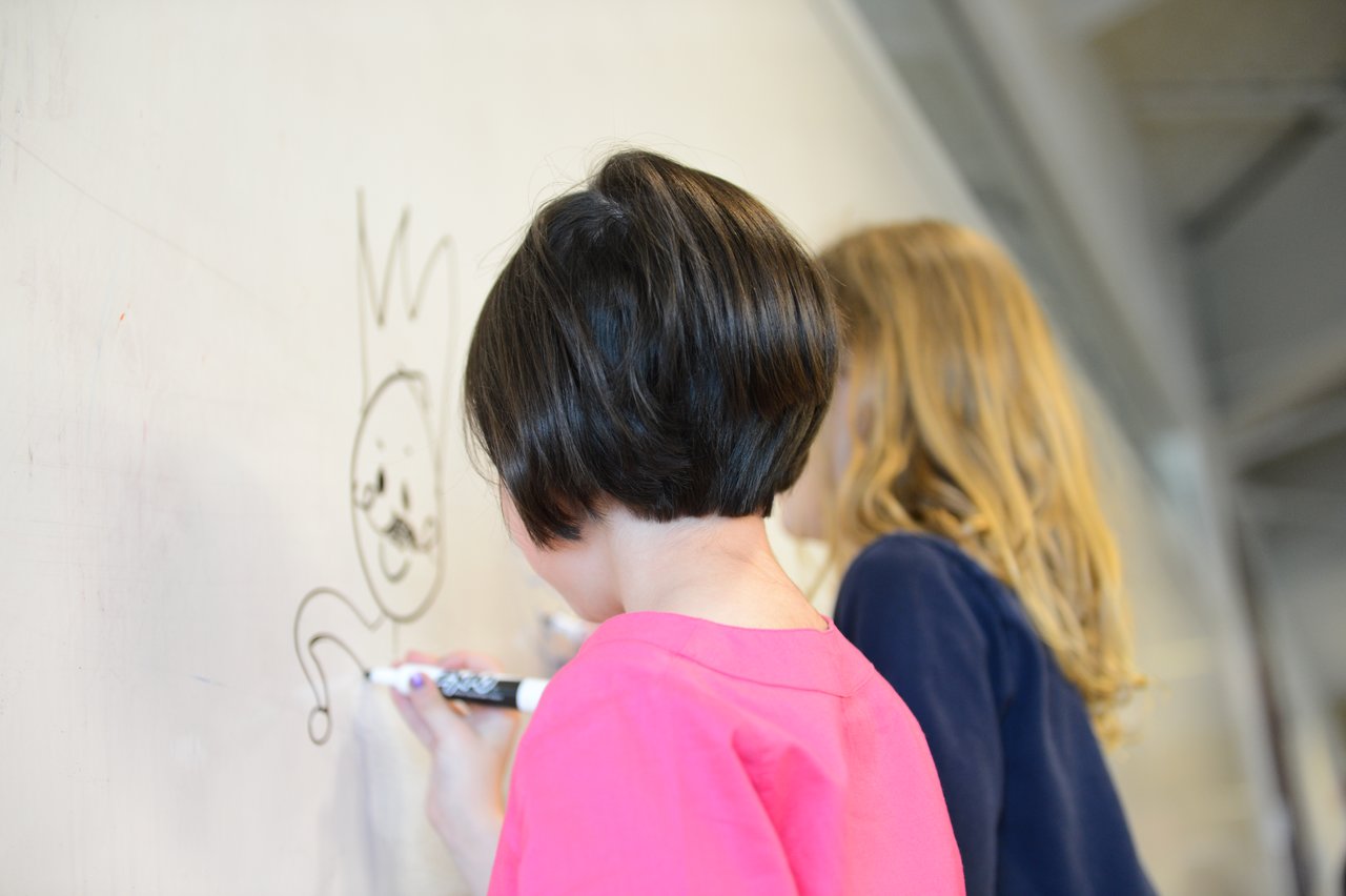 Two children draw on a whiteboard with markers, creating a cartoon-like character with a crown.