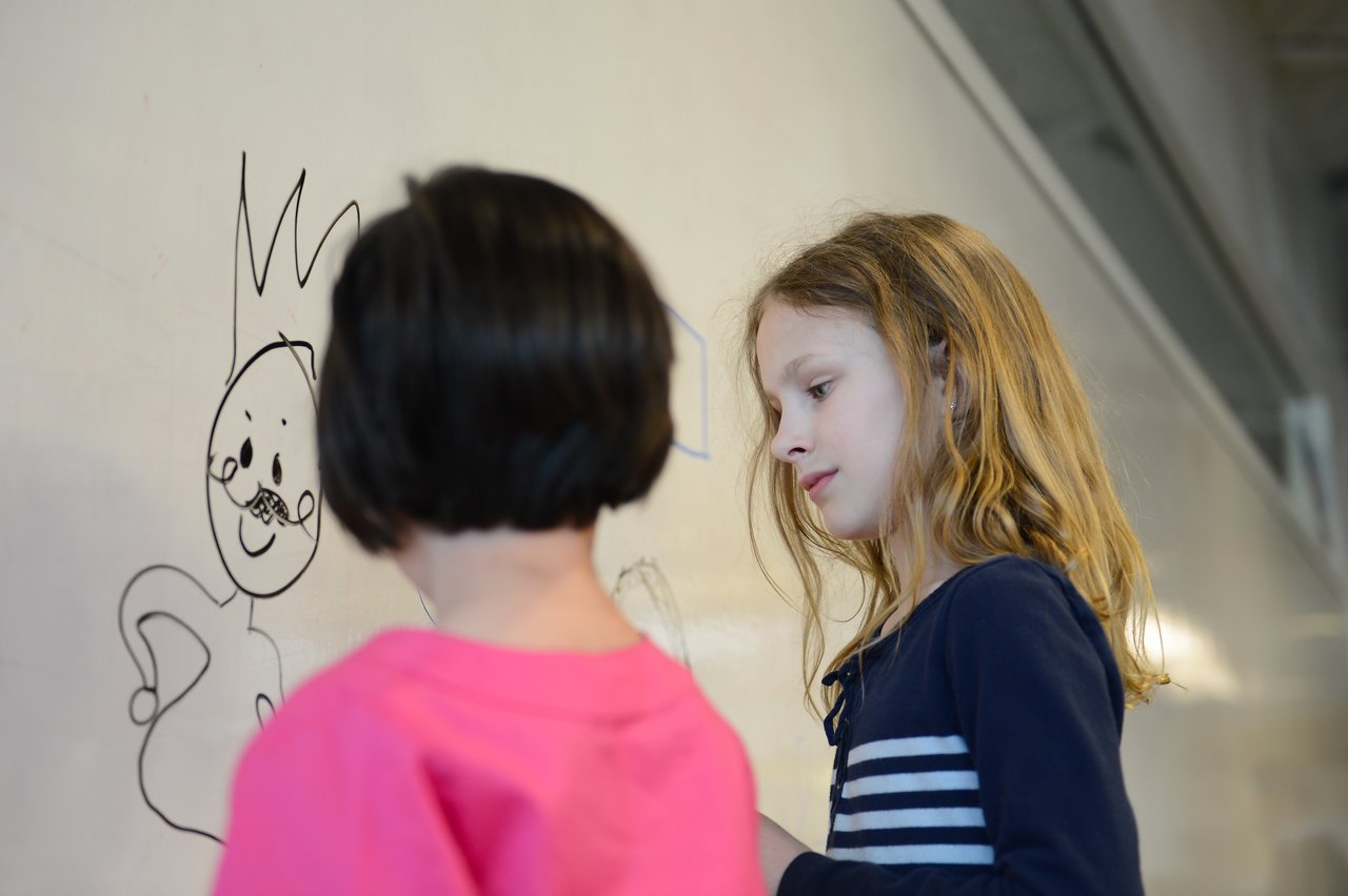Two children draw on a whiteboard, one facing away and the other observing the drawing closely.