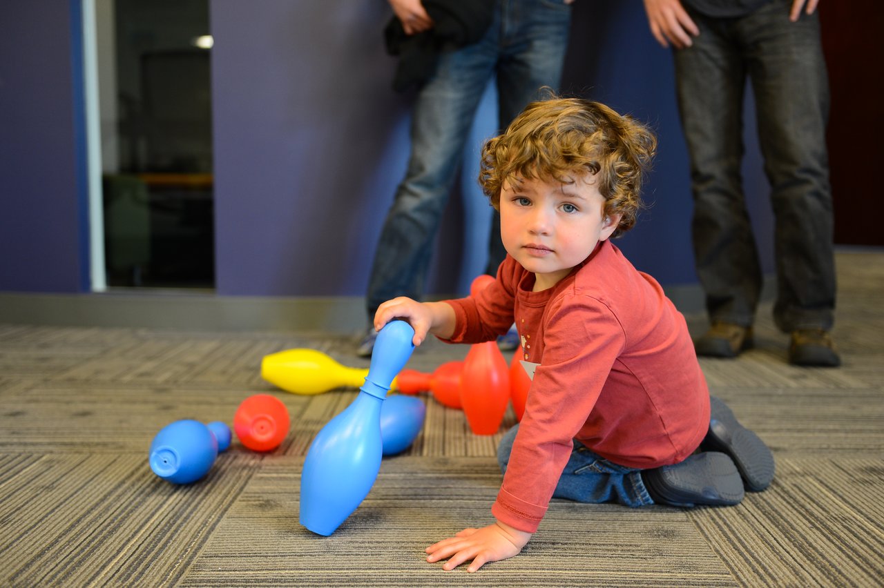 A young child in a red shirt plays with colorful plastic bowling pins on the floor.