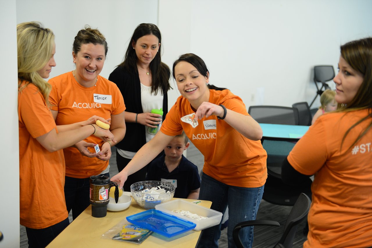 A group of people in orange shirts decorate cookies together at a workplace event with children.