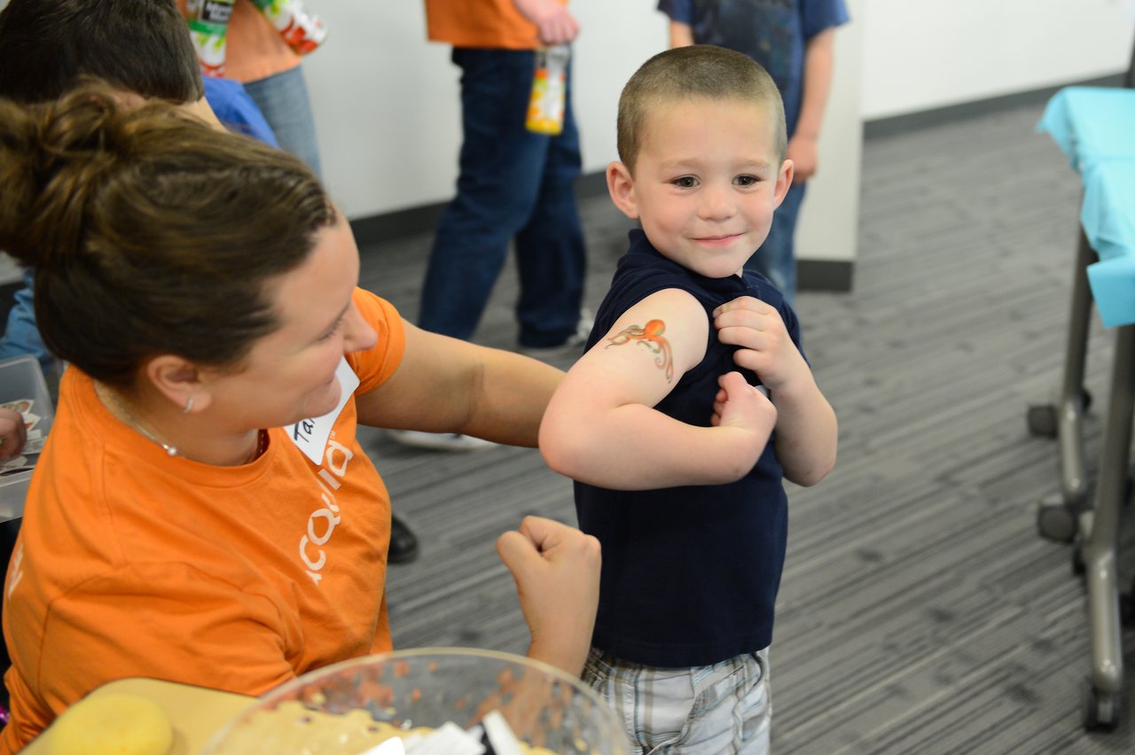 A smiling child flexes their arm to show a temporary tattoo while an adult in an orange shirt looks on.
