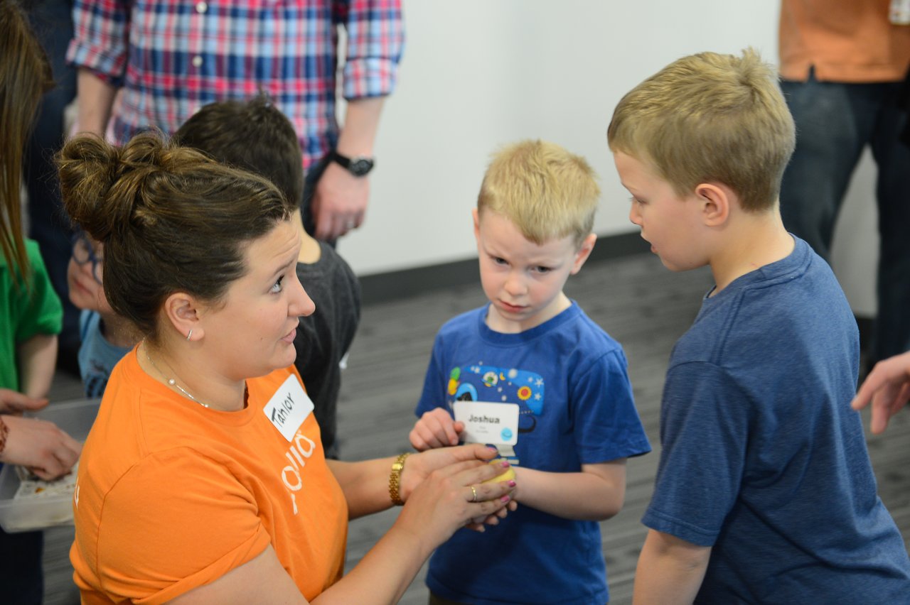 A woman in an orange shirt shows an object to two young boys, who look at it with curiosity.