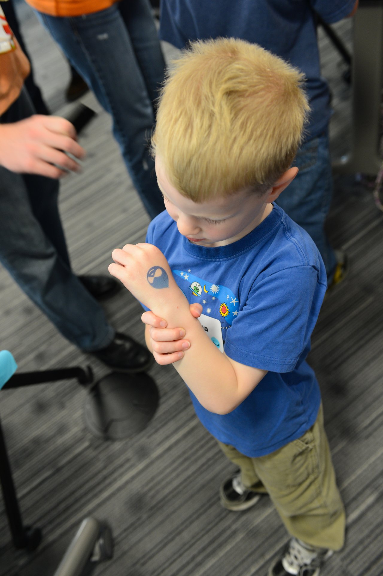 A young child in a blue shirt looks at a temporary tattoo on their arm during an event.