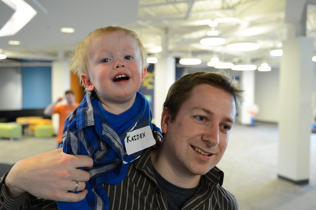 A smiling man carries a young child on his shoulders in an office setting during a family-friendly event.