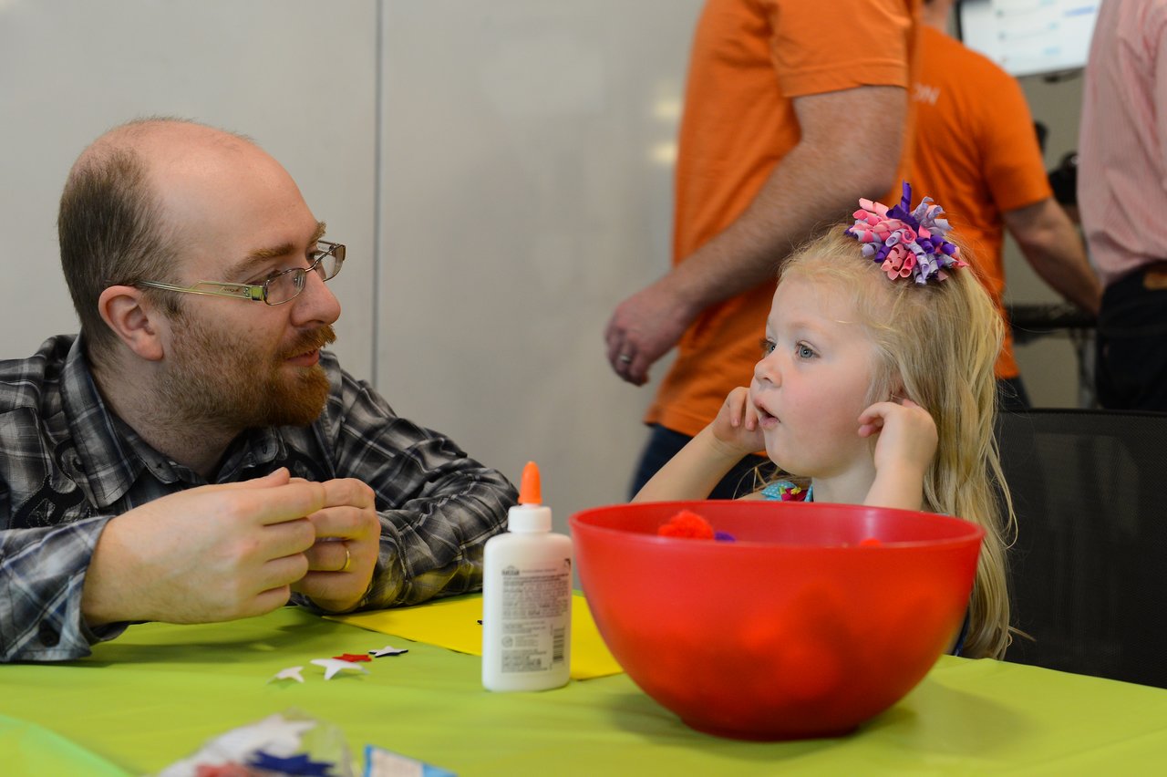 A man and a young girl sit at a table with craft supplies, engaged in conversation.