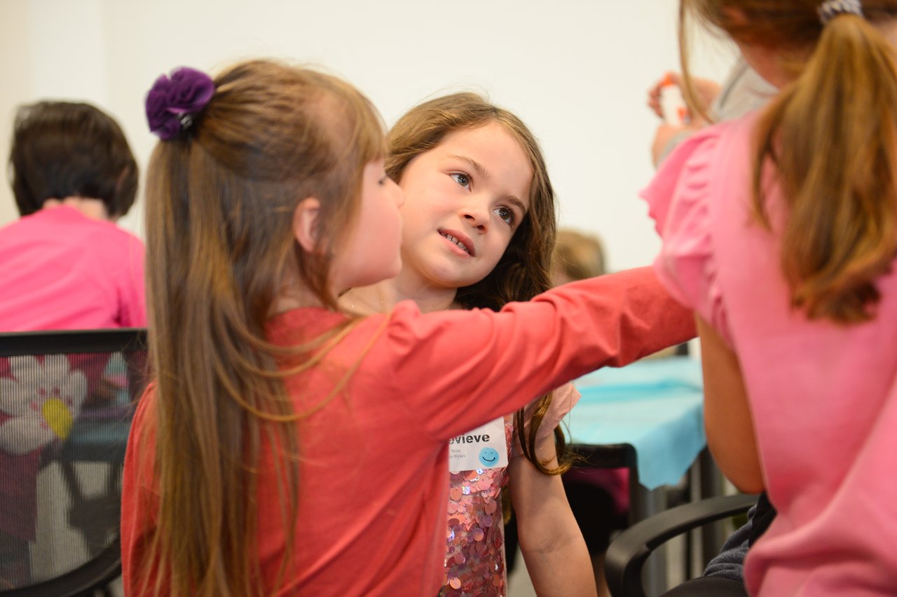 A group of children in pink clothing interact, with one girl reaching toward another in conversation.