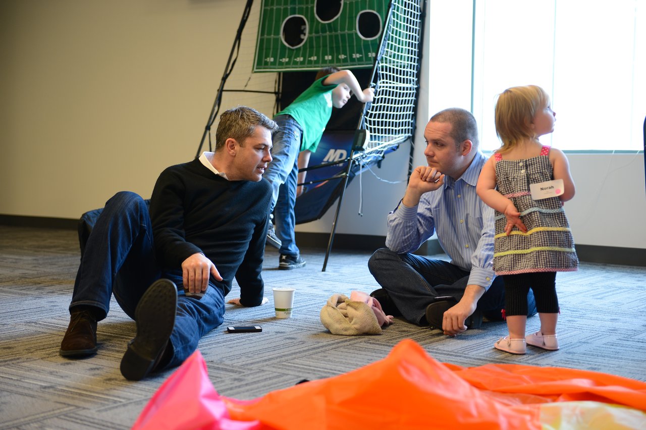 Two men sit on the floor talking while a young girl stands nearby and a boy plays in the background.