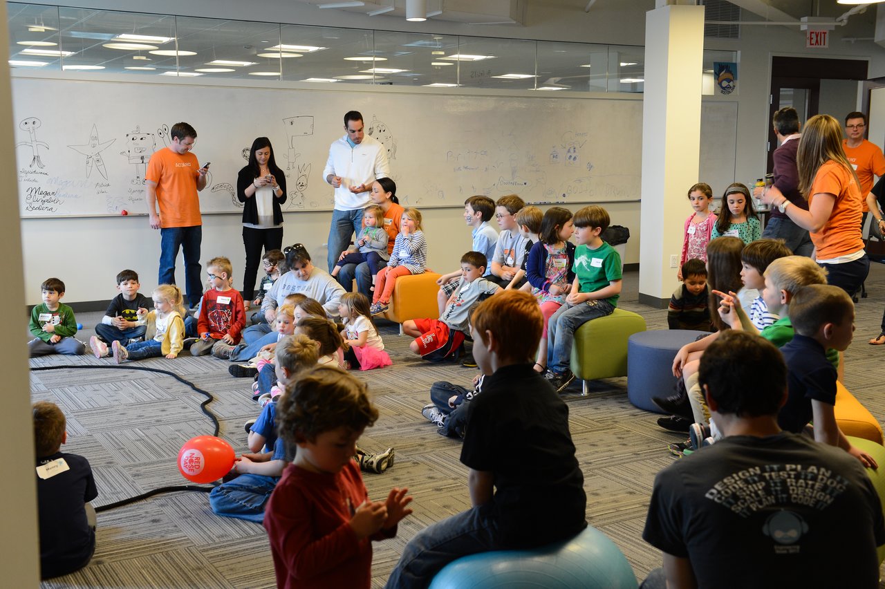 A group of children and adults gather in an office space for a "Bring Your Kids to Work" event.