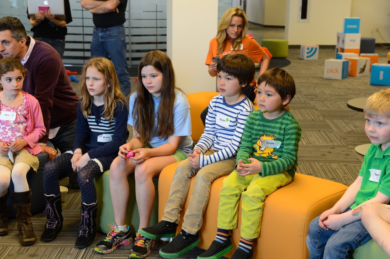 A group of children sits on colorful seats, watching something attentively during a "Bring Your Kids to Work" event.