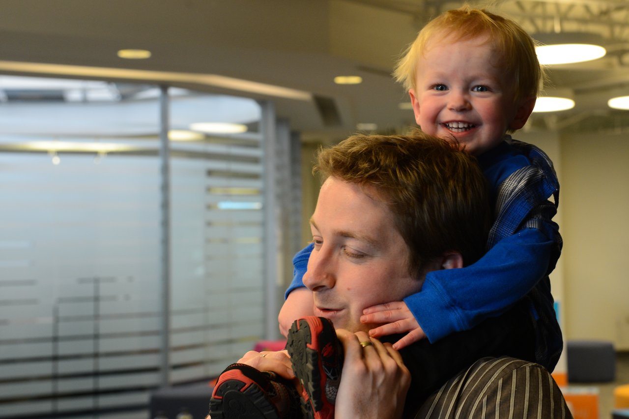 A smiling child in a blue sweater sits on an adult's shoulders, playfully holding onto their face.