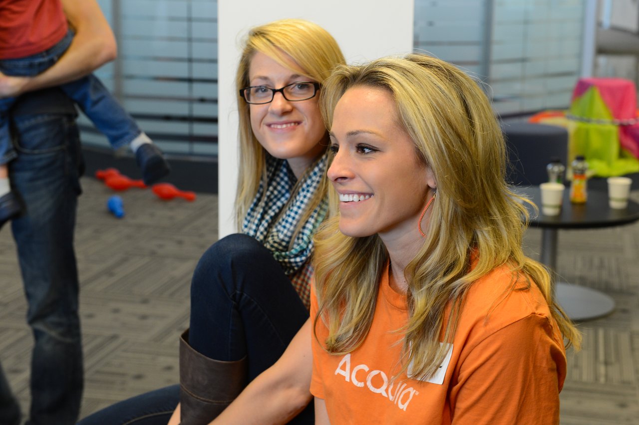 Two smiling women sit together in an office setting, one wearing an Acquia shirt during a company event.