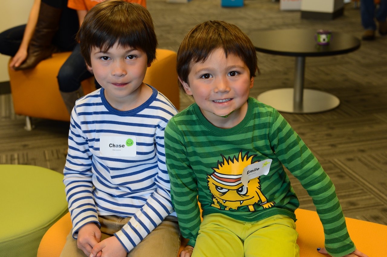 Two young children with name tags sit together on a bench, smiling at the camera during a workplace event.
