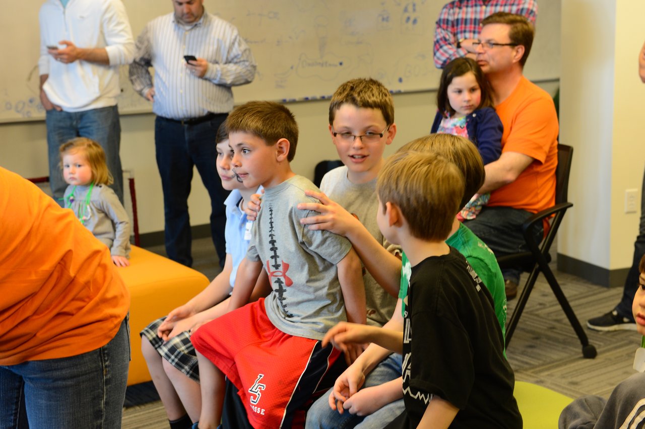 A group of children sits together, talking and interacting, while adults and other children observe in the background.