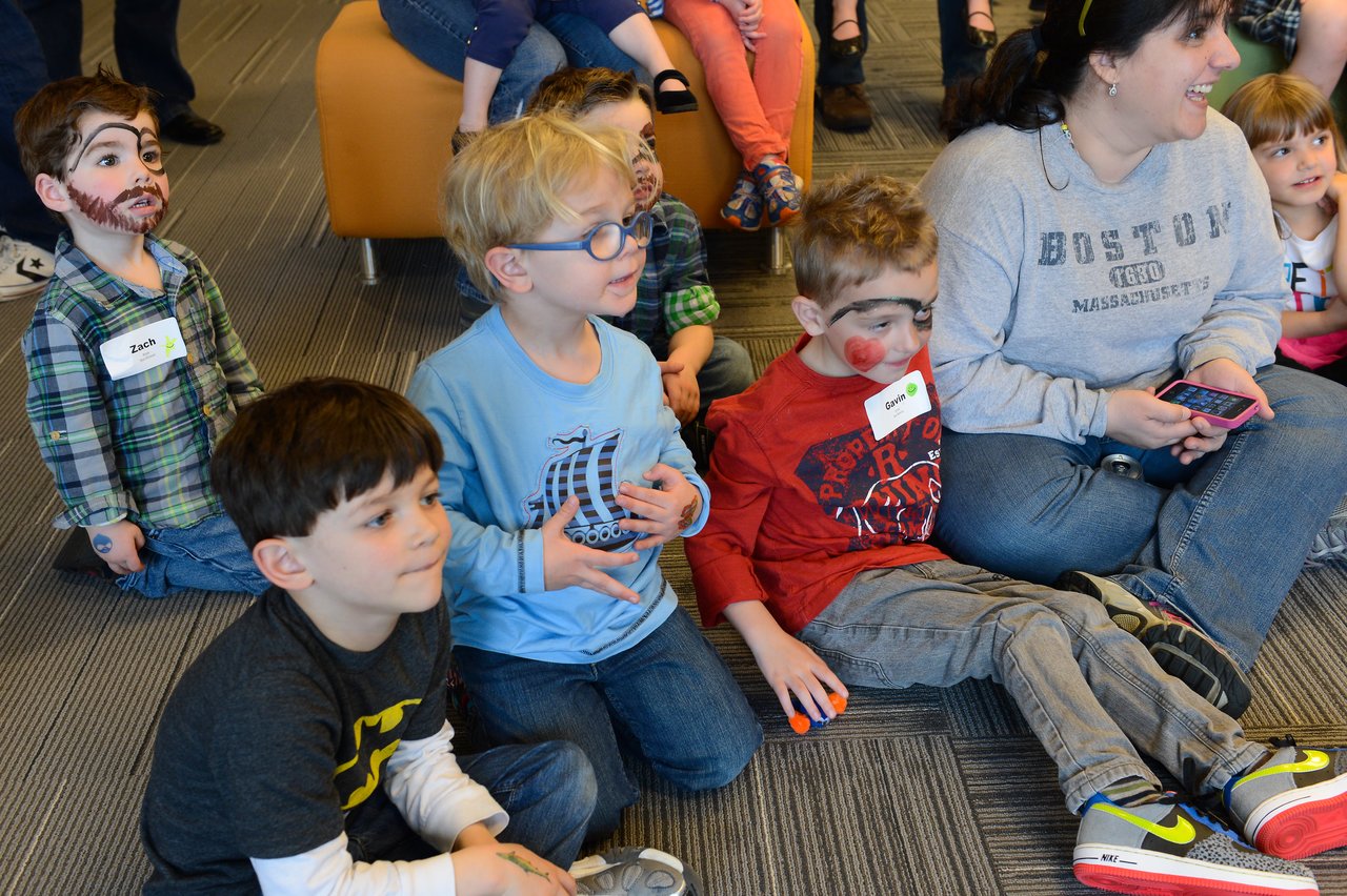 A group of children with face paint sit on the floor, watching something, while an adult sits nearby.