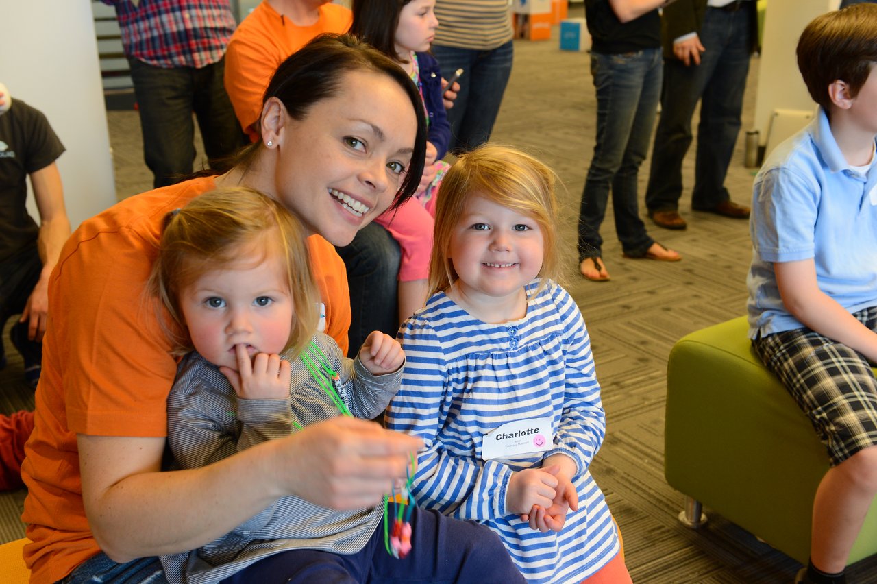 A smiling woman in an orange shirt sits with two young children, one holding beads, at a workplace event.