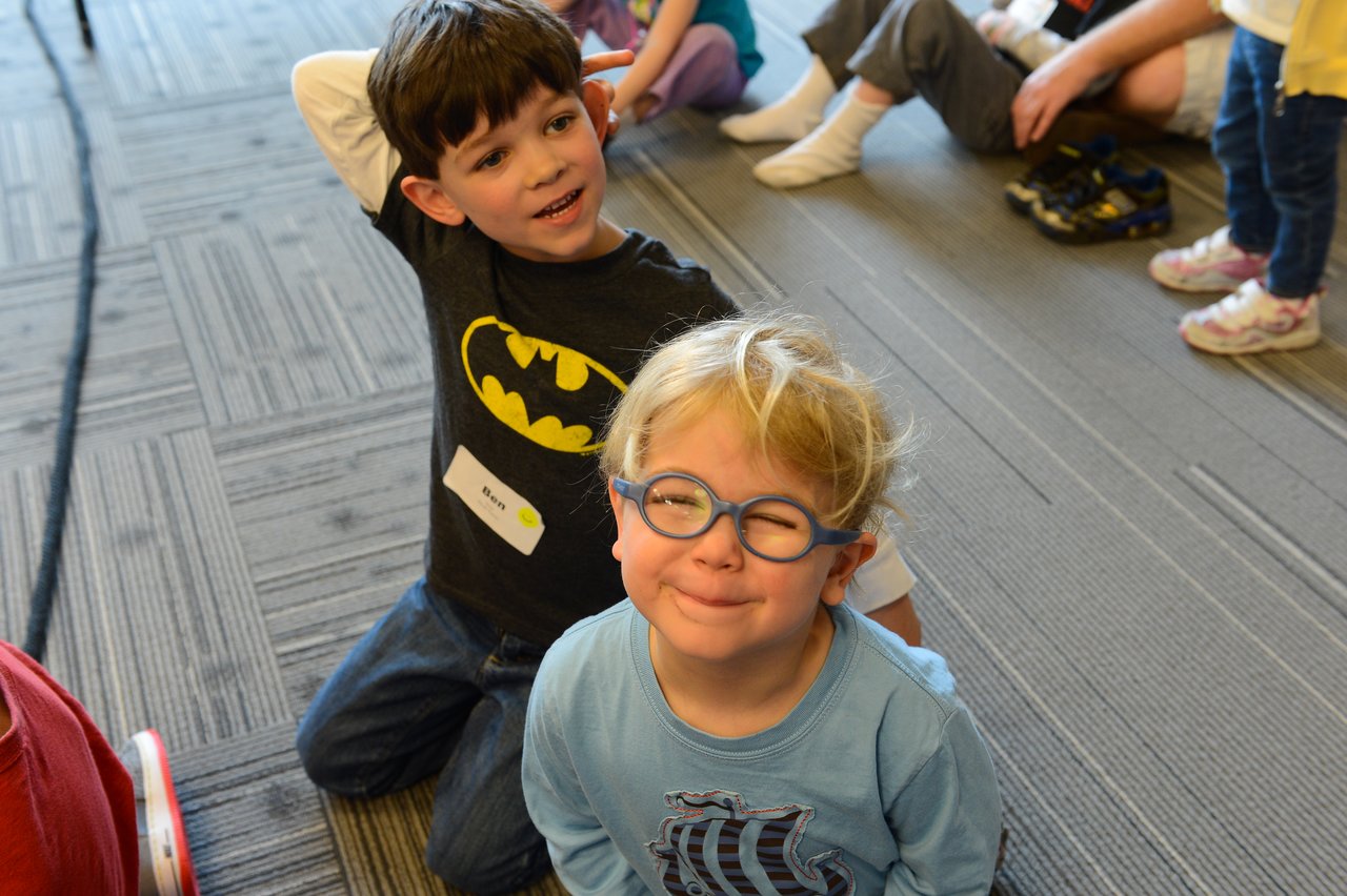 Two young children smile at the camera while sitting on the floor during a workplace event.