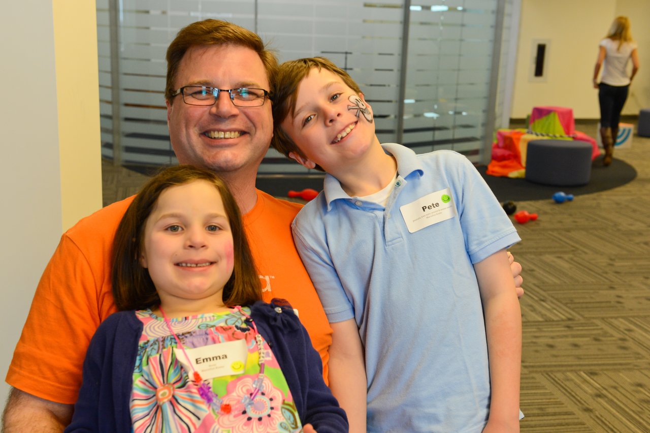 A smiling man sits with two children, one with face paint, at a workplace event.