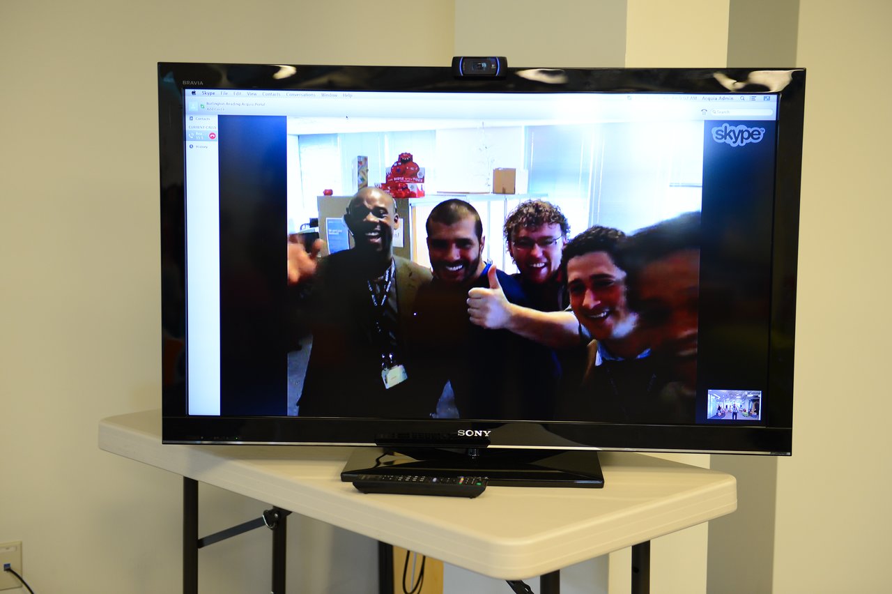 A television screen displays a video call with five smiling people waving and giving a thumbs-up.