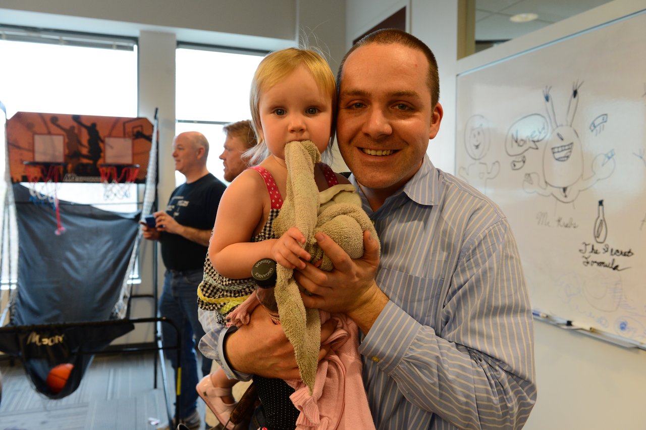 A smiling man holds a young child with a stuffed toy in an office setting.