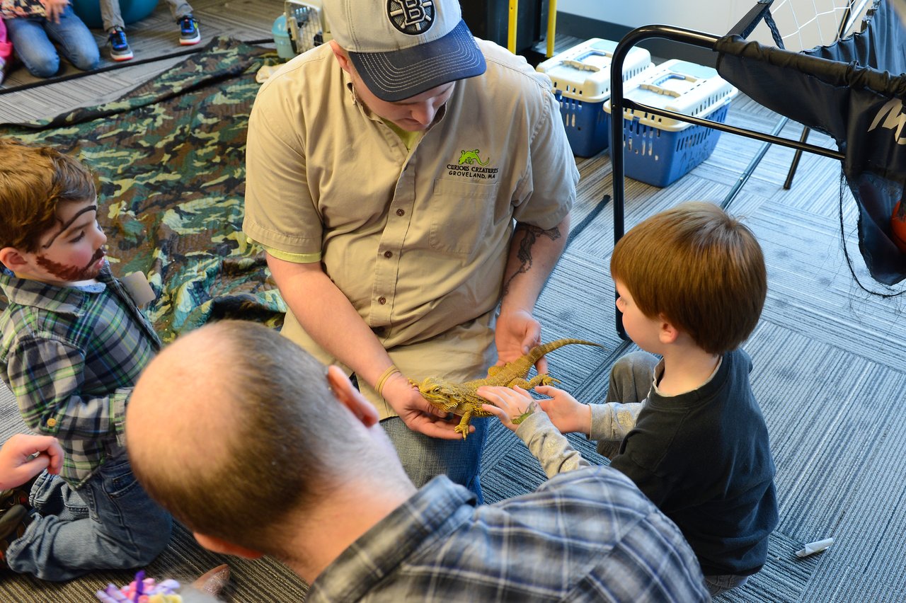 A man in a uniform shows a lizard to a young boy, who reaches out to touch it.