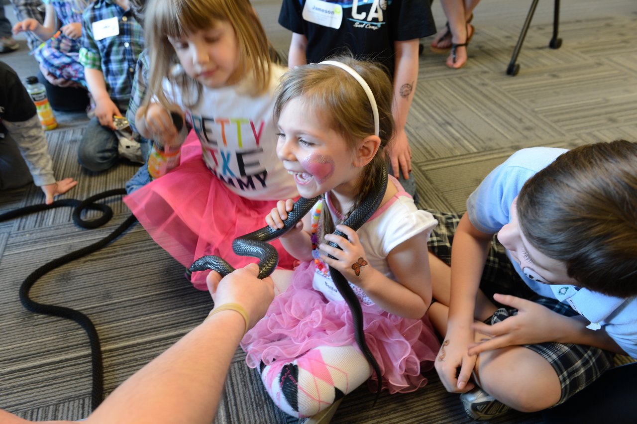 A smiling young girl holds a black snake around her neck while other children watch closely.