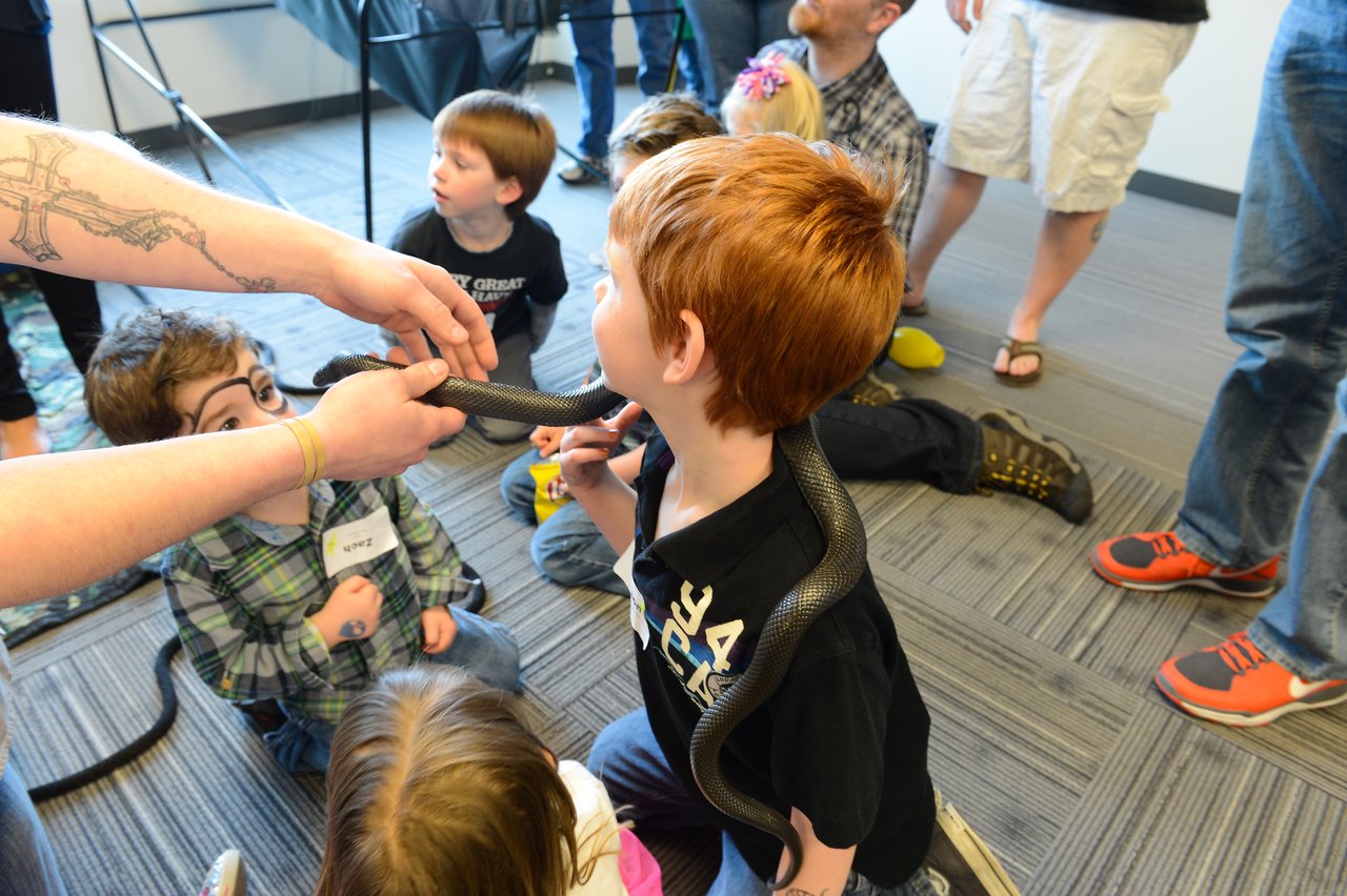 A child with red hair has a snake draped around their shoulders while an adult holds its head.