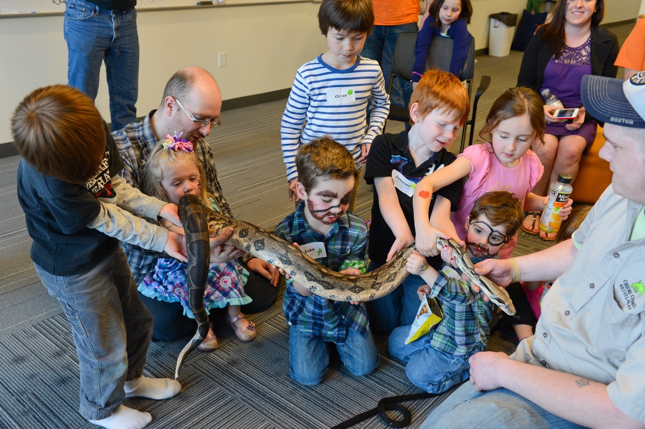A group of children excitedly touch a large snake held by an animal handler during a workplace event.