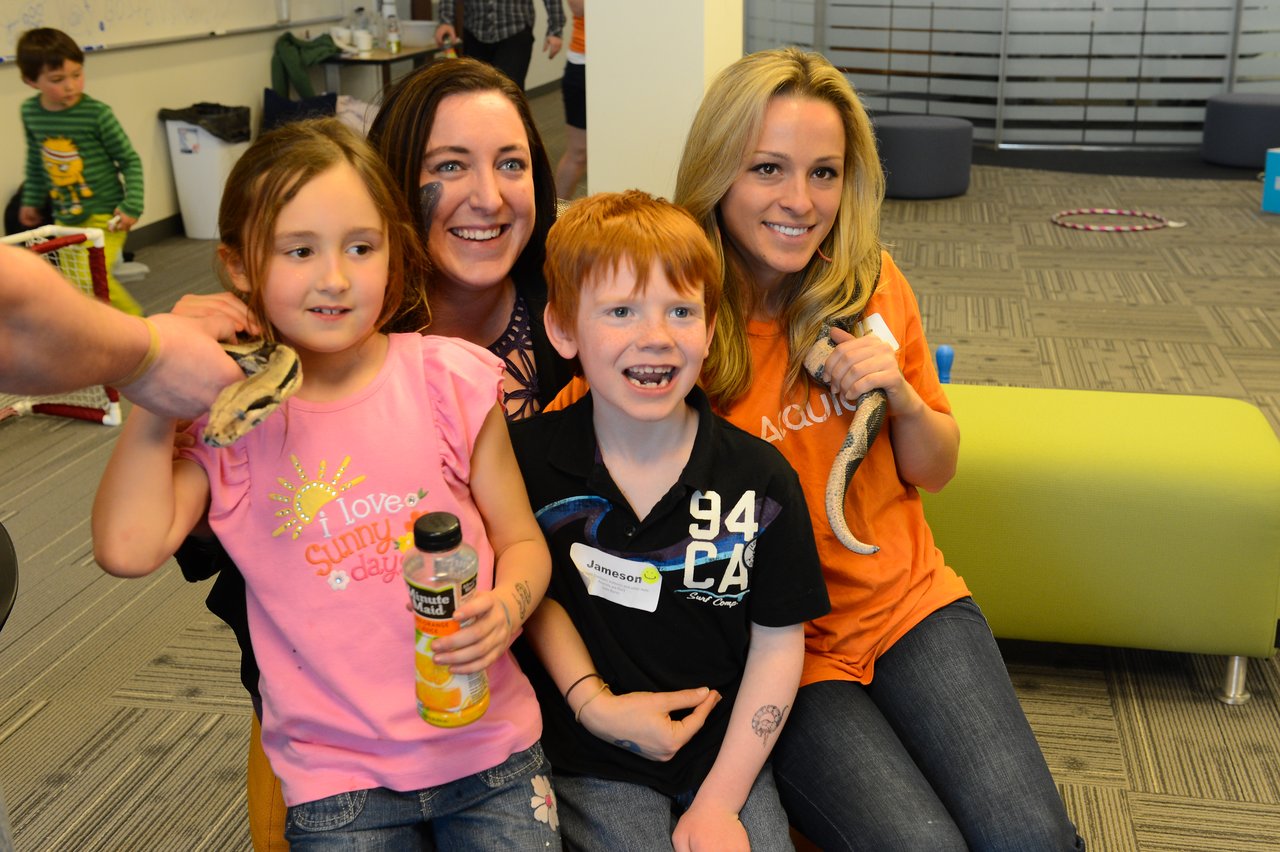 Two women and two children smile while holding snakes at a "Bring Your Kids to Work Day" event.