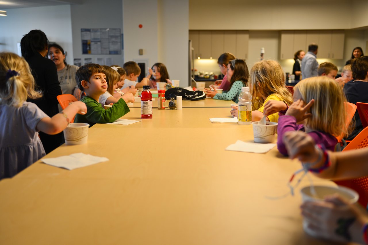 Children sit around a long table, eating and talking during a "Bring Your Kids to Work Day" event.