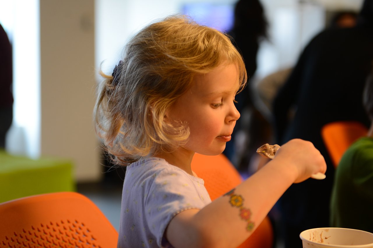 A young girl with a flower tattoo on her arm eats ice cream with a spoon in a busy room.