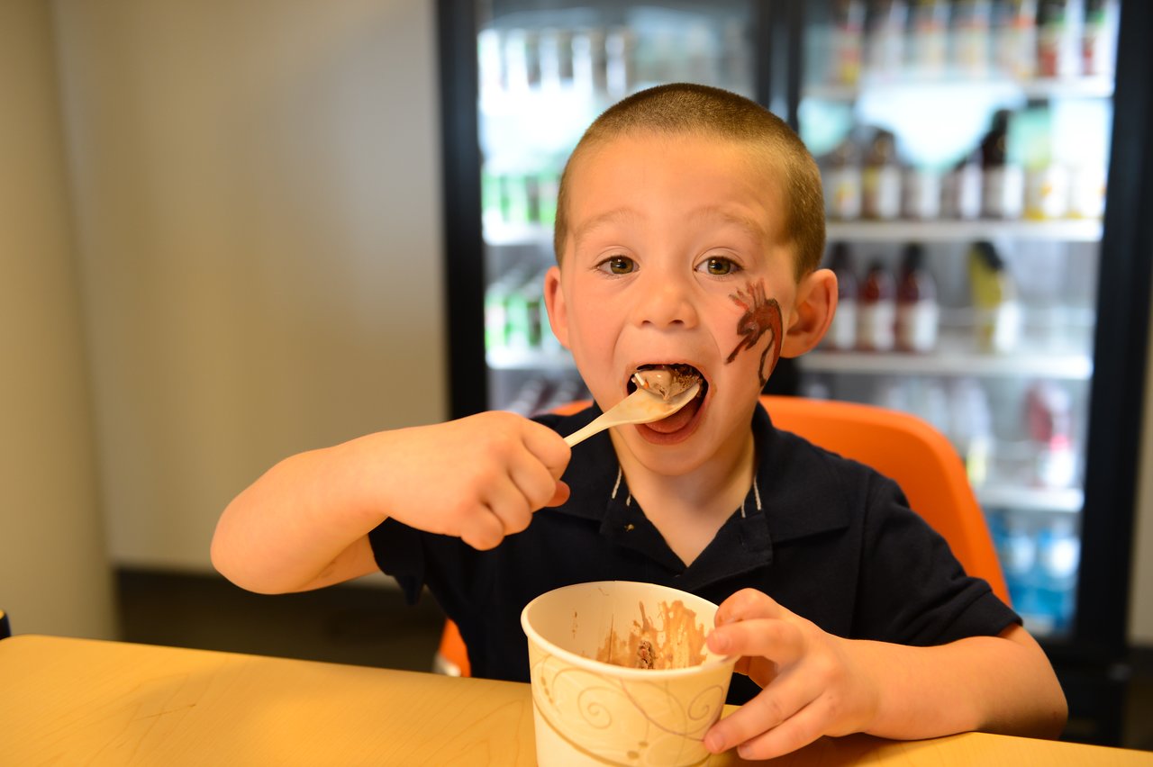 A young child with face paint eats chocolate ice cream with a wooden spoon at a table.