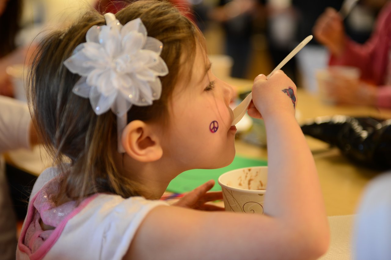 A young girl with a flower hair accessory eats from a cup with a spoon at a gathering.
