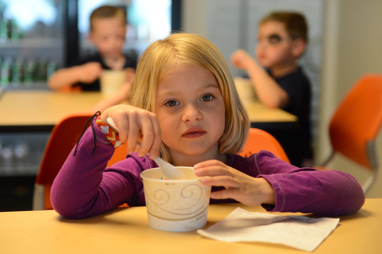 A young girl in a purple shirt eats from a cup with a spoon, sitting at a table.