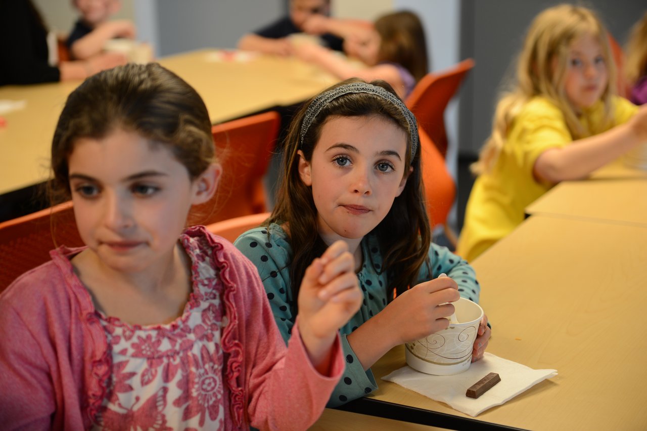 Two young girls sit at a table, one eating from a cup while the other looks away.