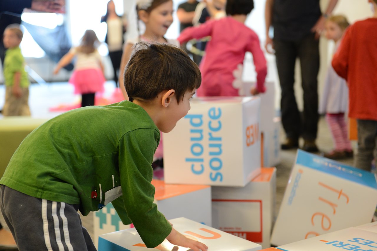 A young child in a green shirt leans forward while playing with large branded boxes at a workplace event.